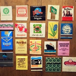 May include: A collection of vintage matchbooks with various designs and text. The matchbooks feature a variety of themes, including travel, hotels, restaurants, and entertainment. Some of the matchbooks have colourful illustrations, while others have simple text designs. The matchbooks are arranged in a grid pattern on a wooden surface.