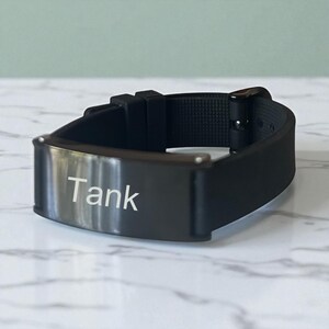 May include: A black wristband with the name "Tank" engraved in white on a metal plate. The band is made of a flexible material and has a buckle closure. The wristband is displayed on a white marble surface.