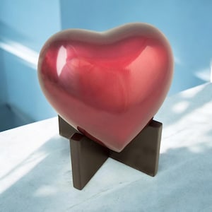 May include: A heart-shaped cremation urn made of polished metal with a deep red finish. The urn is displayed on a dark brown stand.