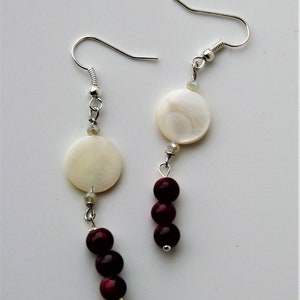 May include: A pair of silver earrings with white and burgundy beads. The earrings feature a round white shell bead and three burgundy beads.