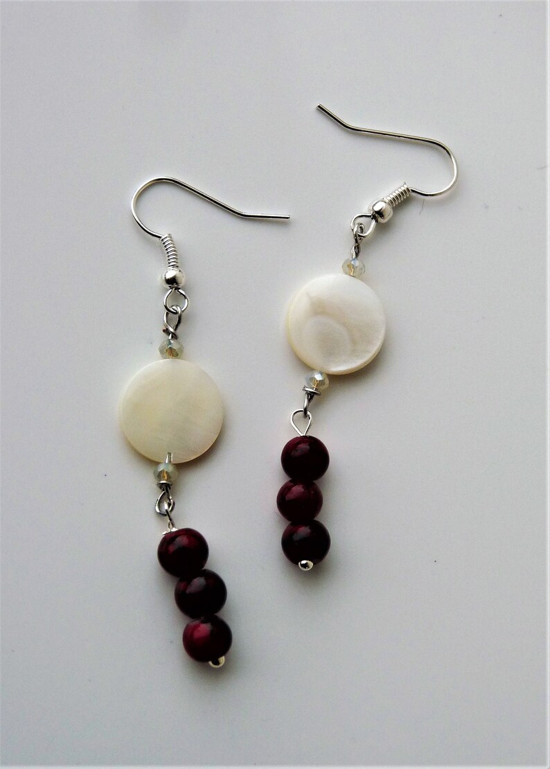 May include: A pair of silver earrings with white and burgundy beads. The earrings feature a round white shell bead and three burgundy beads.