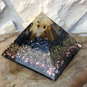 Puede incluir: Una pirámide de cristal de forma piramidal negra con acentos dorados y cobrizos. La pirámide está hecha de resina transparente y contiene varios cristales, incluyendo ojo de tigre, cuarzo transparente y obsidiana negra.