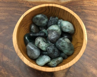 May include: A wooden bowl filled with polished emerald stones. The stones vary in shades of green, from deep forest to pale, with a smooth, rounded shape. The bowl is made of light-colored wood, and the background is a dark wood surface.