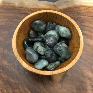 May include: A wooden bowl filled with polished emerald stones. The stones vary in shades of green, from deep forest to pale, with a smooth, rounded shape. The bowl is made of light-colored wood, and the background is a dark wood surface.