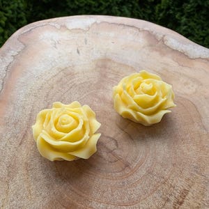 May include: Two yellow rose-shaped objects are displayed on a wooden surface. The roses have detailed petals and a soft, creamy yellow color. The wood grain of the surface is visible, adding a natural element to the composition.