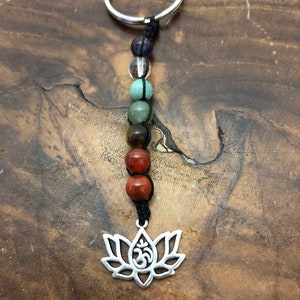 May include: A beaded keyring with a silver lotus flower charm and the Om symbol. The keyring features a series of colourful gemstone beads in shades of purple, clear, green, brown, and red, strung on black cord, and attached to a silver ring.