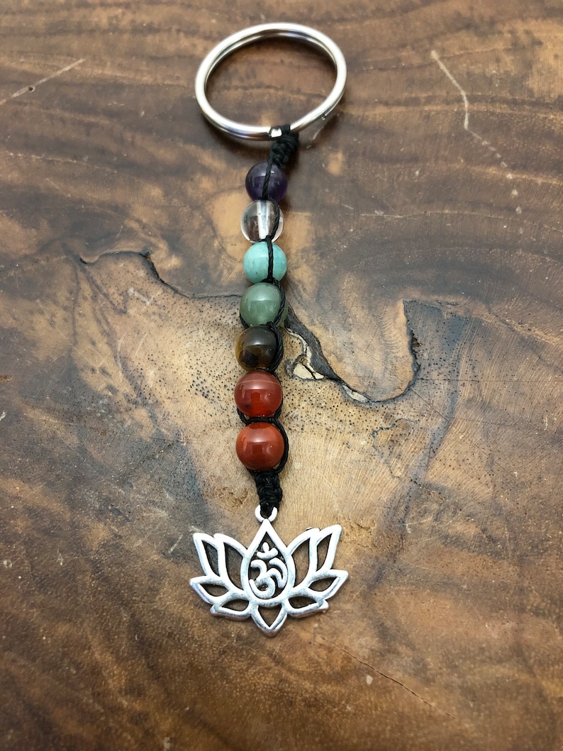 May include: A beaded keyring with a silver lotus flower charm and the Om symbol. The keyring features a series of colourful gemstone beads in shades of purple, clear, green, brown, and red, strung on black cord, and attached to a silver ring.