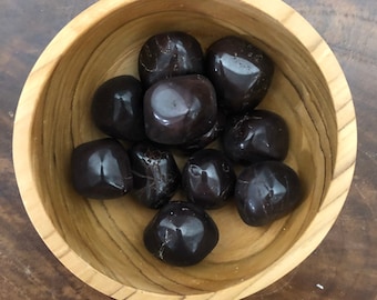 May include: A wooden bowl filled with dark red tumbled stones. The stones are smooth and polished, and they have a natural, earthy look.