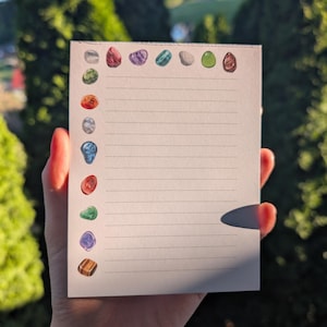 May include: A lined notepad with colorful gemstone illustrations along the left side and top. The gemstones are various colors, including red, purple, green, and brown. The notepad is held in a hand against a blurred green background.