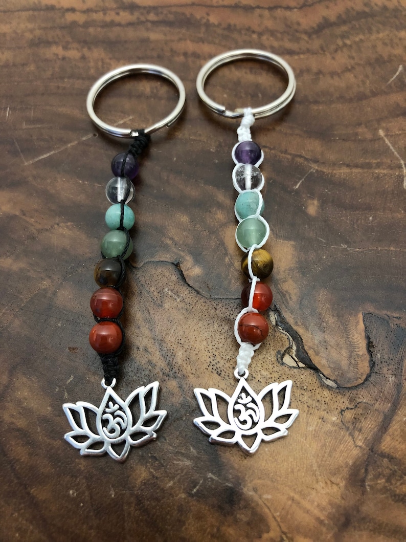 May include: Two keychains featuring silver lotus flower charms and colourful gemstone beads. The beads are arranged in a sequence of colours, and the charms have the Om symbol. The keychains are on a wooden surface.