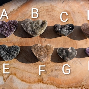 Könnte beinhalten: Acht herzförmige Amethyst- und Quarz-Kristallcluster in verschiedenen Farben, darunter Lila, Grau und Braun. Die Kristalle werden auf einer Holzoberfläche präsentiert, wobei jedes Herz mit einem Buchstaben von A bis H beschriftet ist.