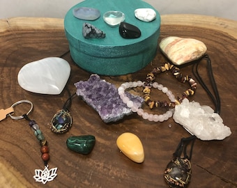 May include: A teal round box with a variety of crystals and stones inside. The box is surrounded by other crystals, stones, and jewelry on a wooden surface.