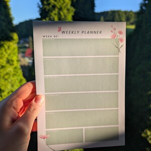 May include: A weekly planner with a light green background and floral accents. The planner has sections for each day of the week, with the text "WEEKLY PLANNER" at the top. A hand is holding the planner.