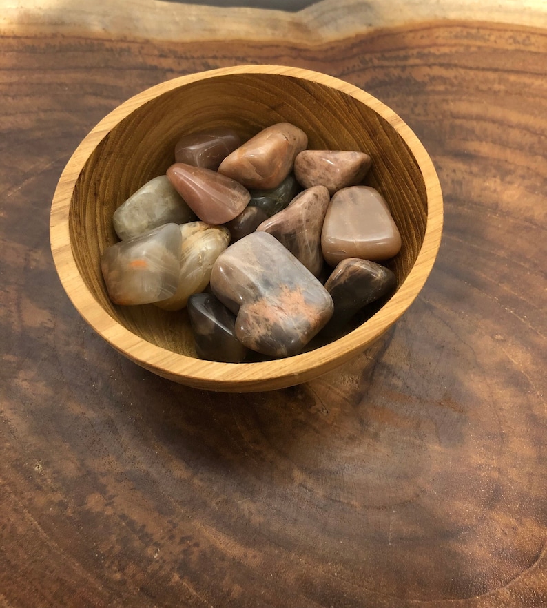 May include: A wooden bowl filled with sunstone tumbled stones. The stones are various shades of pink, orange, and brown.