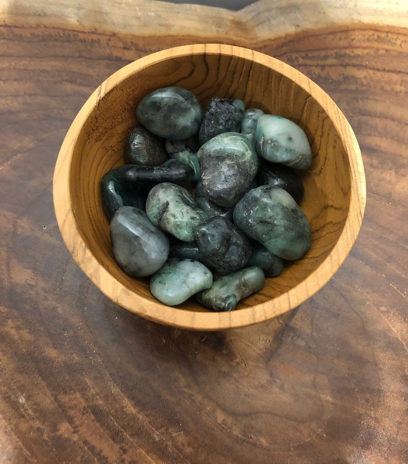 May include: A wooden bowl filled with polished emerald stones. The stones vary in shades of green, from deep forest to pale, with a smooth, rounded shape. The bowl is made of light-colored wood, and the background is a dark wood surface.