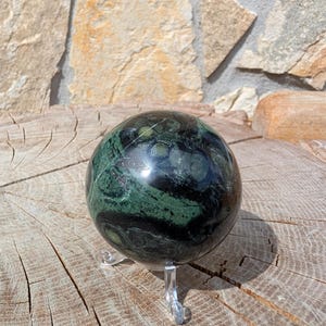 May include: A dark green sphere with black veining, presented on a clear acrylic stand. The polished gemstone ball is set against a natural wood surface and a stone wall background. The sphere's diameter is approximately 8 cm.