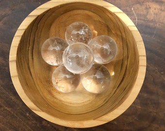 May include: Six clear quartz crystal spheres in a wooden bowl. The bowl is made of light brown wood and has a smooth, polished finish.