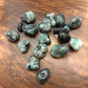 May include: A collection of polished emerald gemstones in various shades of green, with some displaying black inclusions. The stones are smooth and rounded, resting on a wooden surface. The gemstones vary in size and shape.