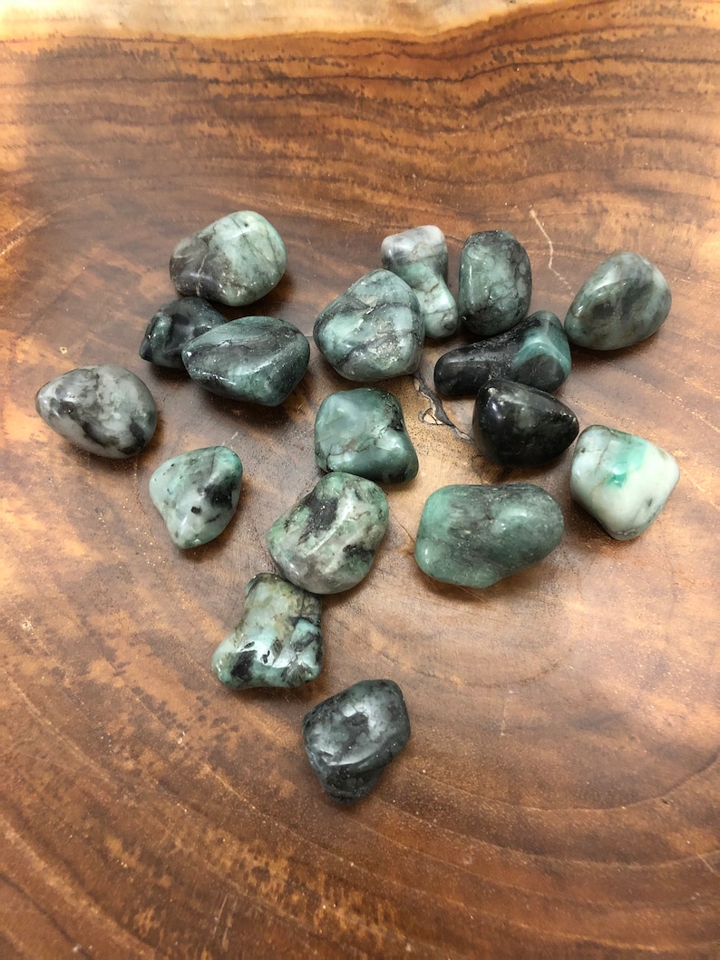 May include: A collection of polished emerald gemstones in various shades of green, with some displaying black inclusions. The stones are smooth and rounded, resting on a wooden surface. The gemstones vary in size and shape.