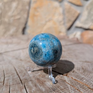 May include: A blue and grey sphere, possibly a decorative stone or crystal ball, is displayed on a clear plastic stand. The sphere has a marbled appearance with shades of blue and grey. The background features a stone wall and a wooden surface.