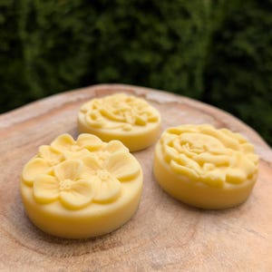 May include: Three round, yellow floral-shaped soaps are arranged on a wooden surface. The soaps have detailed flower designs, with one featuring multiple small flowers. The background is a blurred green.