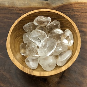 May include: A wooden bowl filled with clear quartz tumbled stones. The stones are smooth and polished, and they have a natural, translucent appearance.