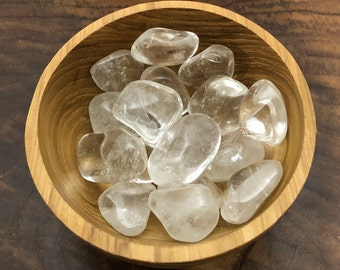 May include: A wooden bowl filled with clear quartz tumbled stones. The stones are smooth and polished, and they have a natural, translucent appearance.