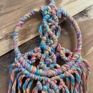 Puede incluir: Un tapiz de macram&eacute; con forma de signo de paz de colores con hilo azul, amarillo y rosa. El tapiz tiene una franja en la parte inferior.
