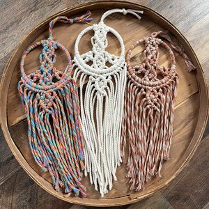 Puede incluir: Tres tapices de macram&eacute; con forma de signo de paz en una bandeja de madera. Uno es blanco, otro es multicolor y el &uacute;ltimo es naranja y marr&oacute;n.