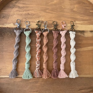 May include: Seven colorful macrame keychains with a twisted rope design and tassels. The keychains are in shades of gray, green, orange, brown, pink, and white.