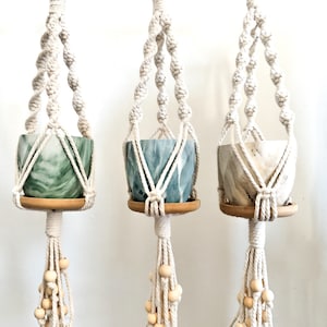 May include: Three macrame plant hangers with wooden rings and white cotton cord. Each hanger has a small wooden base and a tassel with wooden beads. The pots are green, blue, and white with marbleized patterns.