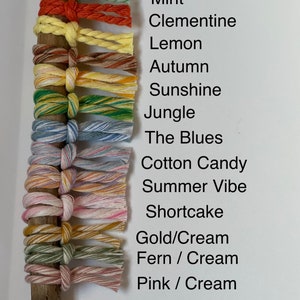 Puede incluir: Primer plano de una varilla de madera con 14 colores diferentes de cord&oacute;n envueltos alrededor. Los cordones est&aacute;n etiquetados con nombres como "Menta", "Clementina", "Lim&oacute;n", "Oto&ntilde;o", "Sol", "Jungla", "Los Blues", "Algod&oacute;n de az&uacute;car", "Ambiente de verano", "Pastel de queso", "Oro/Crema", "Helecho/Crema" y "Rosa/Crema".