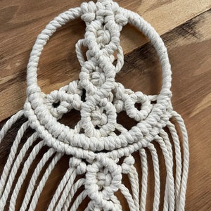 Puede incluir: Un atrapasue&ntilde;os de macram&eacute; blanco en forma de signo de paz con flecos. El signo de paz est&aacute; hecho de cuerda anudada y tiene una forma circular.