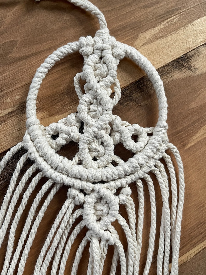 Puede incluir: Un atrapasue&ntilde;os de macram&eacute; blanco en forma de signo de paz con flecos. El signo de paz est&aacute; hecho de cuerda anudada y tiene una forma circular.