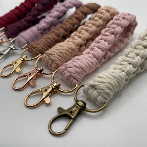 May include: A set of five colorful macrame keychains with gold and silver key rings. The keychains are made with a variety of colors, including pink, brown, and white.