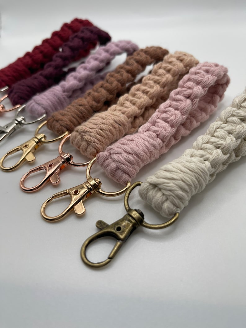 May include: A set of five colorful macrame keychains with gold and silver key rings. The keychains are made with a variety of colors, including pink, brown, and white.