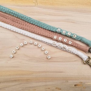 May include: Three macrame lanyards in white, dusty rose, and teal, each with a name spelled out in wooden letter beads. The word "PERSONALIZE" is spelled out with individual beads. Each lanyard has a bronze-colored clasp.