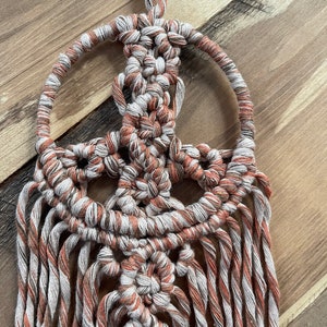 Puede incluir: Un tapiz de macram&eacute; de pared con un dise&ntilde;o de signo de paz. El tapiz est&aacute; hecho con hilo blanco y naranja y tiene flecos largos.