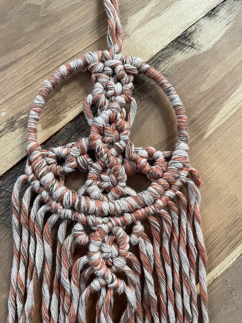 Puede incluir: Un tapiz de macram&eacute; de pared con un dise&ntilde;o de signo de paz. El tapiz est&aacute; hecho con hilo blanco y naranja y tiene flecos largos.
