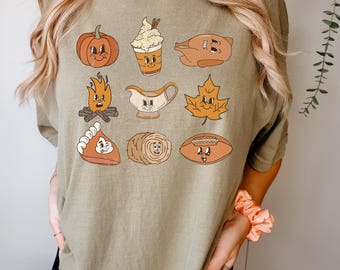 May include: Khaki-colored t-shirt featuring a collection of cartoon illustrations of Thanksgiving-themed items. The graphics include a pumpkin, a coffee drink, a turkey, a campfire, a gravy boat, a leaf, a pie, a hay bale, and a football.