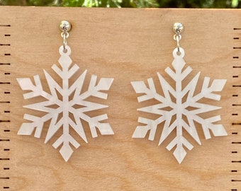 Op de afbeelding: Een paar witte sneeuwvlok oorbellen met zilveren oorhaakjes. De oorbellen zijn ongeveer 2,5 cm in diameter en worden getoond op een houten liniaal met de tekst "Moon Child Trinkets".