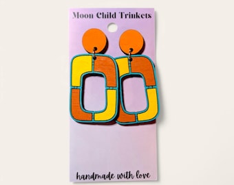 Puede incluir: Pendientes de madera con diseño retro. Los pendientes presentan círculos naranjas en la parte superior y formas rectangulares con detalles amarillos, naranjas y azules. Los pendientes están en una tarjeta morada que dice "Moon Child Trinkets" y "handmade with love."