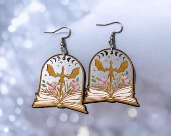Könnte beinhalten: Ein Paar Ohrringe aus Holz mit einem silbernen Drachenmotiv und einem Buch mit Blumen. Die Ohrringe sind in einem silbernen, weißen und rosafarbenen Farbschema bemalt.