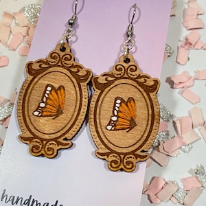 May include: A pair of wooden earrings with a butterfly design in a frame. The earrings are brown with orange and white butterflies. The earrings are handmade and have silver hooks. The card says "Moon Child Trinkets" and "handmade with love".