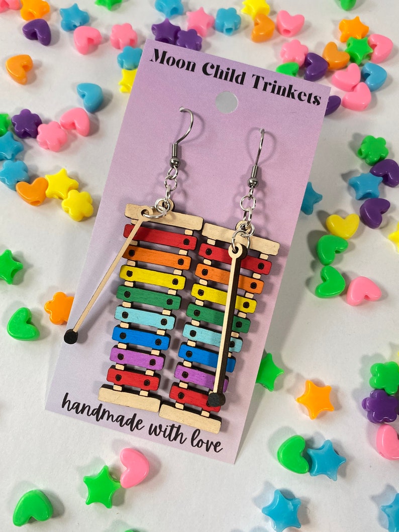 Puede incluir: Pendientes de xil&oacute;fono arco&iacute;ris con ganchos plateados. Los pendientes est&aacute;n hechos de madera y tienen un mazo negro unido a cada uno. Los pendientes est&aacute;n hechos a mano y la tarjeta dice "Moon Child Trinkets" y "hecho con amor".
