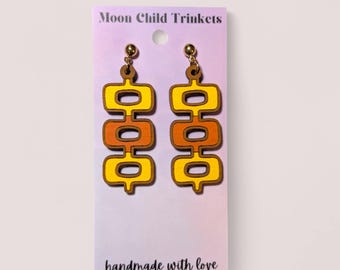 Puede incluir: Un par de pendientes colgantes en una tarjeta de presentación con el texto Moon Child Trinkets y handmade with love. Los pendientes presentan tres formas rectangulares redondeadas apiladas en amarillo y naranja, suspendidas de postes dorados.