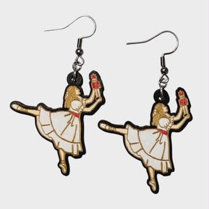 May include: A pair of dangle earrings featuring a ballerina design. Each earring depicts a ballerina in a white dress with a red sash, holding a red object. The earrings have silver-colored hooks and black accents.