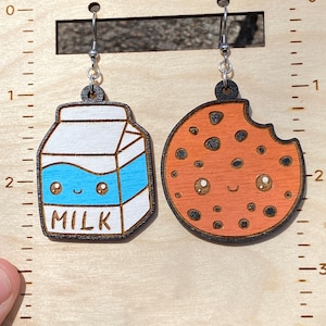 May include: A pair of wooden earrings featuring a milk carton and a chocolate chip cookie. Both charms have cute cartoon faces with eyes and a smile. The earrings are hanging from silver hooks.