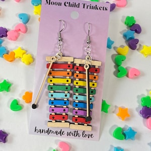 Puede incluir: Pendientes de xil&oacute;fono arco&iacute;ris con ganchos plateados. Los pendientes est&aacute;n hechos de madera y tienen un mazo negro unido a cada uno. La tarjeta dice "Moon Child Trinkets" y "hecho a mano con amor".