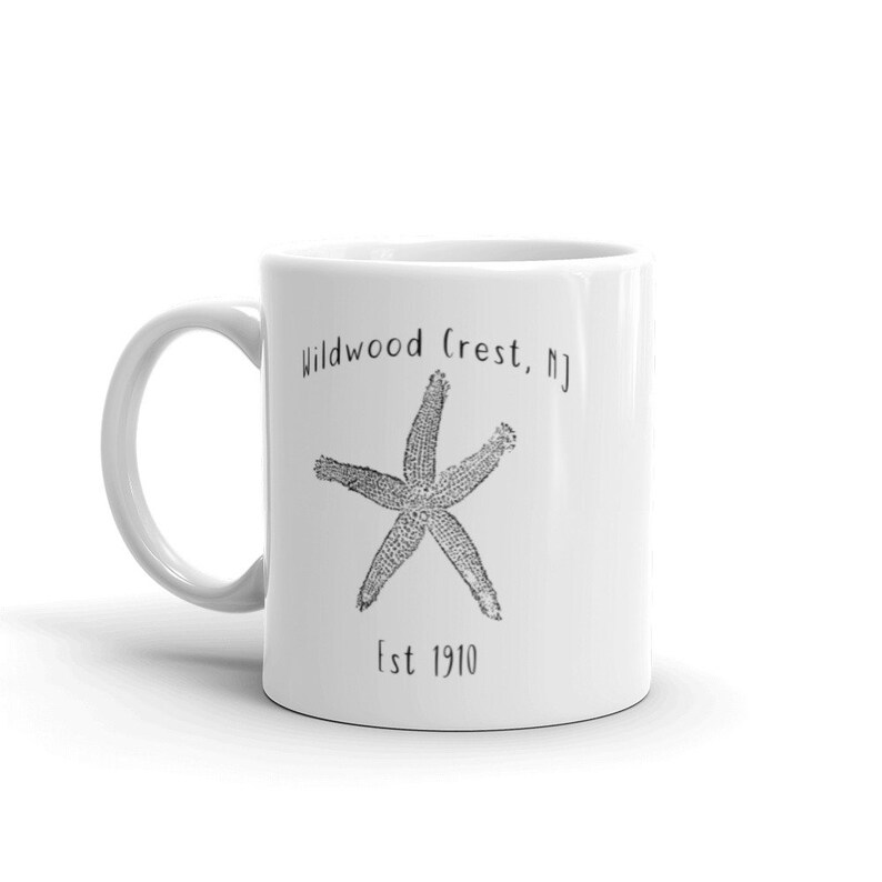 May include: White ceramic mug with a black starfish design and the text "Wildwood Crest, NJ" and "Est 1910". The mug has a curved handle and is suitable for hot or cold beverages.